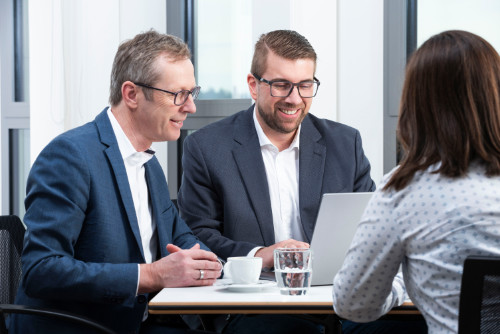 Webdesign Wiesbaden – Persönliche Beratung für digitale Strategien Zwei Berater im Anzug im Gespräch mit einer Kundin an einem Tisch mit Laptop, Wasser und Kaffee. Das Bild zeigt ein Beratungsgespräch zum Thema professionelles Webdesign und digitale Strategien in Wiesbaden.