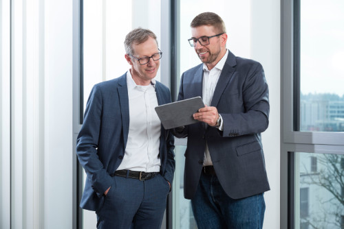 Webdesign Wiesbaden – Strategische Beratung für erfolgreiche Websites Zwei Männer im Business-Outfit stehen in einem hellen Büro und betrachten gemeinsam ein Tablet. Das Bild zeigt eine Beratungssituation zur Planung und Optimierung von Webdesign-Projekten in Wiesbaden.