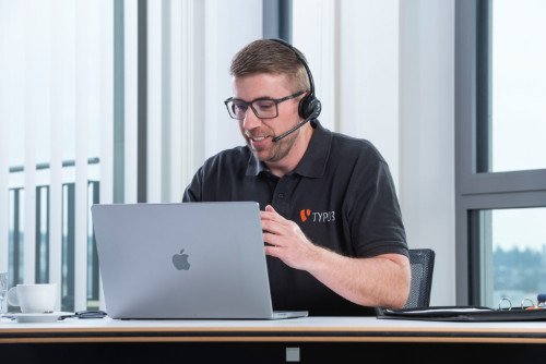 Webdesign Wiesbaden – TYPO3 Experte im Online-Meeting Ein Mann mit Headset sitzt vor einem Laptop und führt ein Online-Meeting. Er trägt ein schwarzes TYPO3-Shirt und spricht mit einem Kunden über Webdesign-Lösungen in Wiesbaden.